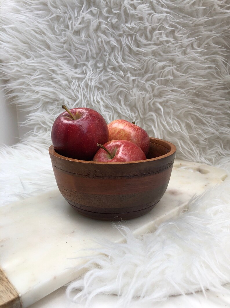 Op de afbeelding: Een houten kom gevuld met drie rode appels. De kom heeft een donkerbruin houtnerfpatroon en staat op een wit, pluche oppervlak.