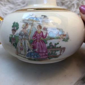 May include: A white ceramic teapot with a gold trim and a painted scene of three women in a garden setting. The women are wearing colorful dresses and holding an umbrella and flowers.