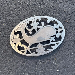 May include: A silver metal whale weathervane with an oval frame and decorative cutouts. The whale is in the center, with an arrow and other nautical-themed designs. The weathervane has a small loop at the top for hanging.