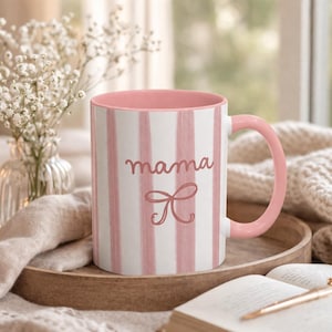 May include: A pink and white striped ceramic mug with the word "mama" in cursive script and a bow graphic. The mug has a pink handle and interior. The mug is on a wooden tray with a book and pen.