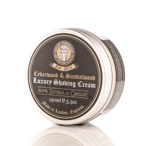 Puede incluir: Una lata plateada con una etiqueta negra que dice "Cedarwood & Sandalwood Luxury Shaving Cream". La etiqueta también incluye un logotipo de un vikingo con un hacha y el texto "Est. 2015". La lata es de 150 ml o 5,3 oz y está hecha en Londres, Inglaterra.