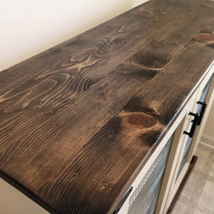 The dark stained planked pine top of a cream Farmhouse Coffee Bar Cabinet, with oil-rubbed bronze bar pulls on its welded wire mesh doors