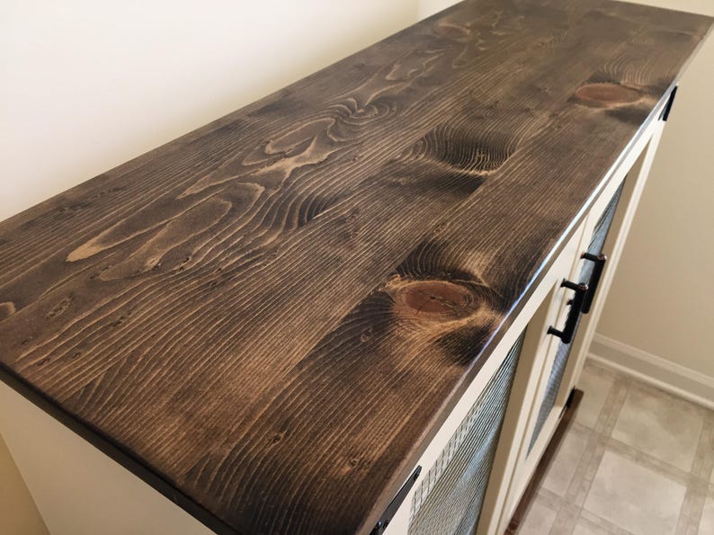 The dark stained planked pine top of a cream Farmhouse Coffee Bar Cabinet, with oil-rubbed bronze bar pulls on its welded wire mesh doors