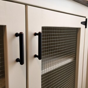 Close view of the cream Farmhouse Coffee Bar Cabinet's inset doors: welded wire mesh panels, oil-rubbed bronze bar pulls and a black T-hinge