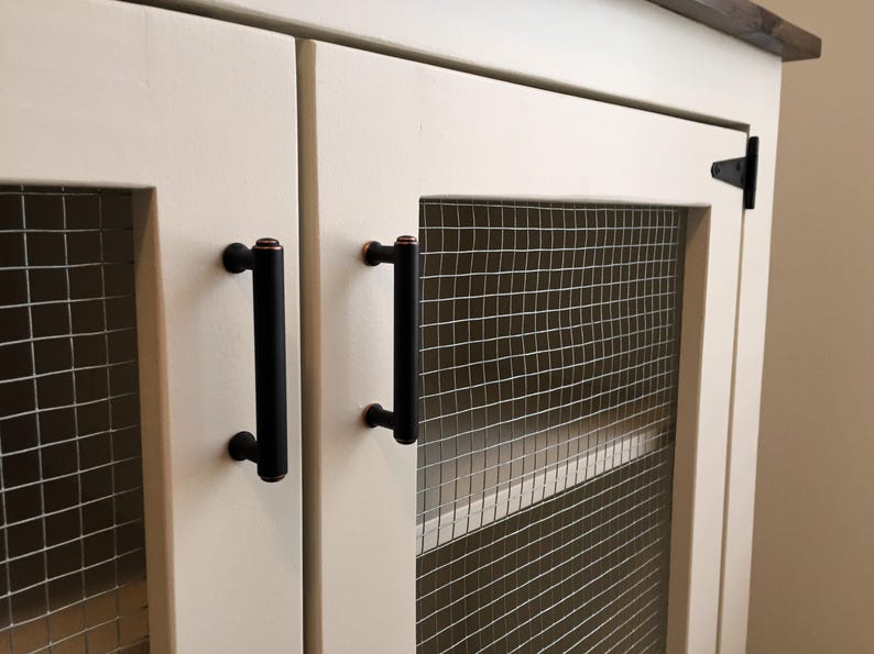 Close view of the cream Farmhouse Coffee Bar Cabinet's inset doors: welded wire mesh panels, oil-rubbed bronze bar pulls and a black T-hinge