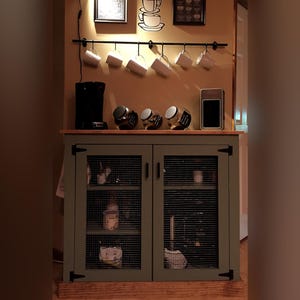 Farmhouse Coffee Bar Cabinet painted sage green in use as a coffee bar: a coffee maker and jars of coffee on top, supplies on the shelves behind the welded wire mesh doors