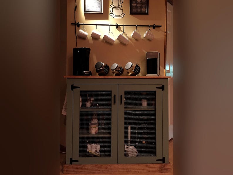 Farmhouse Coffee Bar Cabinet painted sage green in use as a coffee bar: a coffee maker and jars of coffee on top, supplies on the shelves behind the welded wire mesh doors