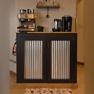 Farmhouse Coffee Bar Cabinet painted black, with corrugated metal doors and a stained top and base trim, set up as a coffee bar with an espresso machine and a coffee maker