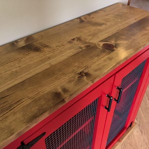 The stained planked pine top of the red Farmhouse Coffee Bar Cabinet, with its bar pulls, a black T-hinge and welded wire mesh doors