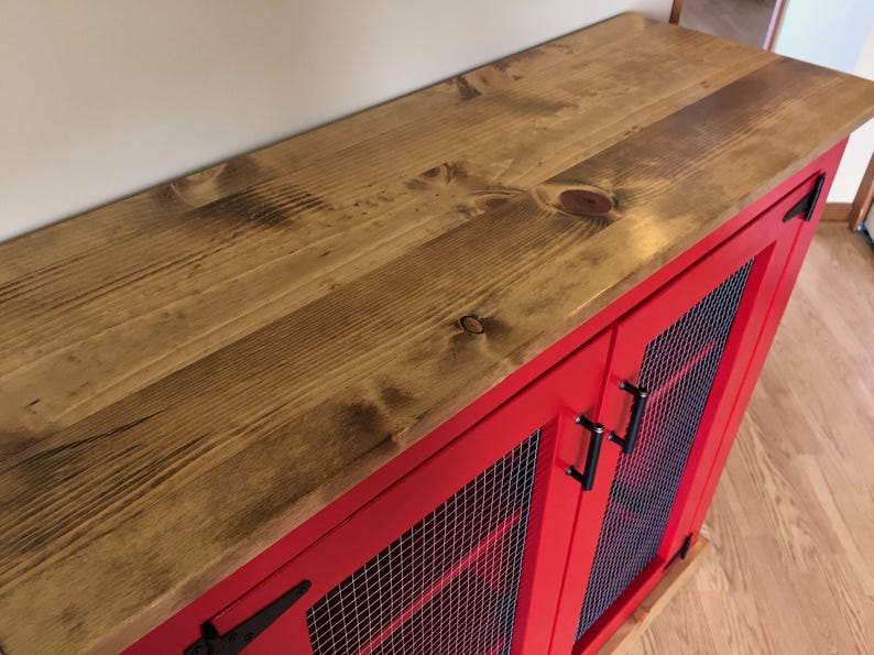 The stained planked pine top of the red Farmhouse Coffee Bar Cabinet, with its bar pulls, a black T-hinge and welded wire mesh doors