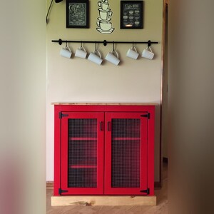 Farmhouse Coffee Bar Cabinet painted red with welded wire mesh doors, set up as a coffee station below a rail of hanging mugs and coffee prints