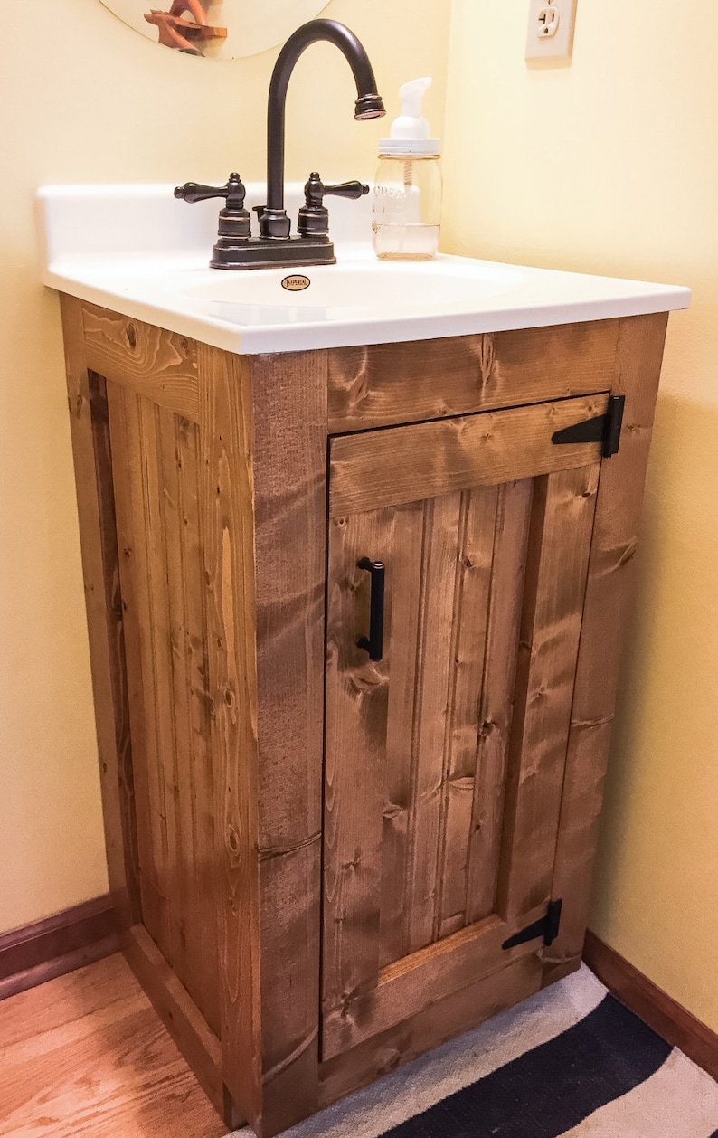 Small rustic bathroom vanity base in a golden stain with oil-rubbed bronze hinges, holding a white vanity sink top