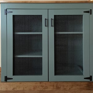 Farmhouse Coffee Bar Cabinet painted sage green, with welded wire mesh doors, black T-hinges, bar pulls and a stained top and base trim; the shelves show through the mesh
