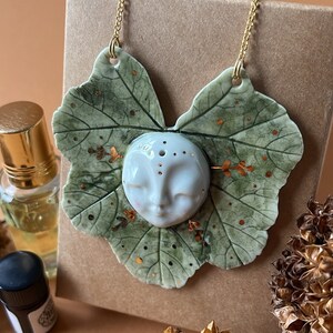 May include: A ceramic necklace with a leaf design and a small face in the center. The necklace is gold and has a green and white leaf design. The face is white with gold accents.