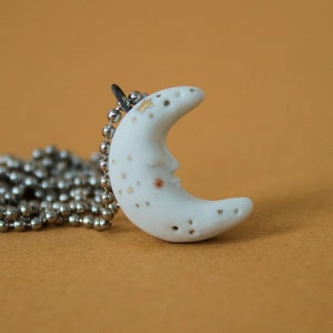 May include: A white ceramic crescent moon pendant with gold flecks hangs on a silver ball chain necklace.