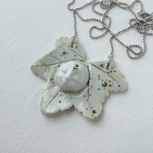 May include: A white porcelain necklace pendant shaped like a leaf with gold stars and a crescent moon design. The pendant is attached to a silver chain.