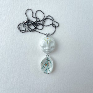 Puede incluir: Un collar de porcelana blanca con un colgante de cara y un colgante de hoja. El colgante de cara tiene detalles verdes y amarillos. El colgante de hoja tiene detalles verdes y marrones. El collar está en una cadena negra.