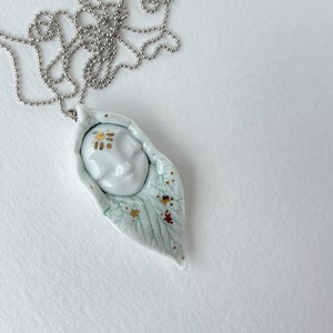 May include: A white porcelain pendant necklace featuring a face with gold accents, set within a leaf-shaped design with green and gold details.