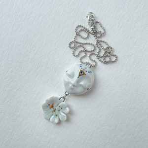 May include: A white ceramic necklace with a pendant featuring a stylized face with blue and gold accents. The pendant is attached to a white ceramic flower with gold accents.