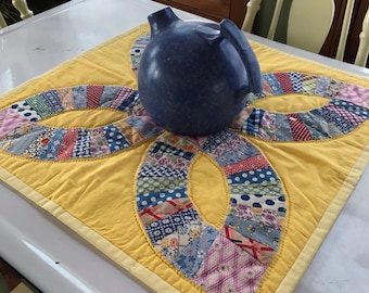 May include: A blue ceramic pitcher sits on a yellow quilted table runner with a patchwork design. The runner has a yellow border and is made of various fabrics with floral and geometric patterns.