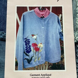 Peut inclure: Un motif d'appliqué de vêtement bleu clair avec un colibri, des fleurs et le texte "Hummm... Bug" et "Stitchin' Sisters".