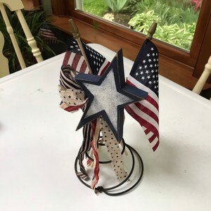 May include: A patriotic centerpiece featuring miniature American flags, a distressed wooden star, and decorative ribbons. The star is painted white with a navy blue border. The flags and ribbons have red, white, and blue patterns.