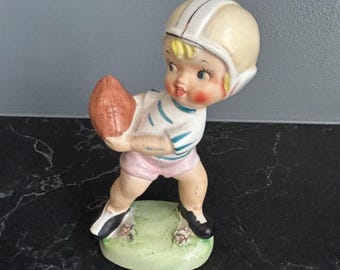 May include: A vintage ceramic figurine of a child in a football uniform, holding a brown football. The figurine wears a white helmet with stripes, a white and blue striped shirt, pink shorts, and black shoes. The base is green.