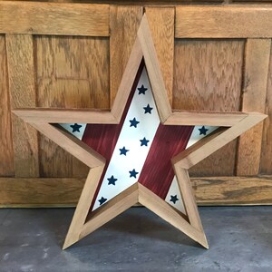 May include: A wooden star-shaped frame with a red, white and blue American flag design inside. The frame is made of natural wood and has a rustic look.