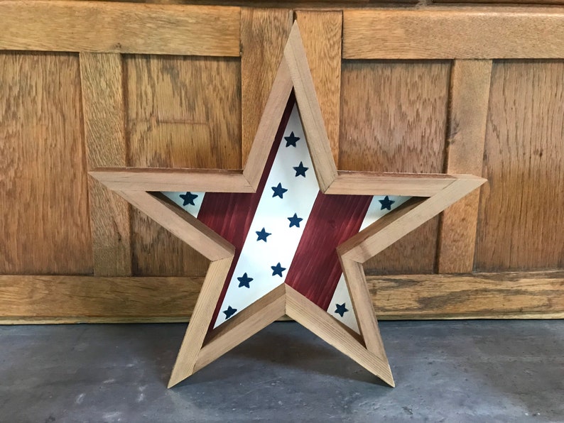 May include: A wooden star-shaped frame with a red, white and blue American flag design inside. The frame is made of natural wood and has a rustic look.