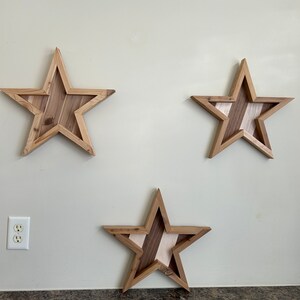 May include: Three wooden stars with a striped pattern in shades of brown and white, mounted on a light beige wall.