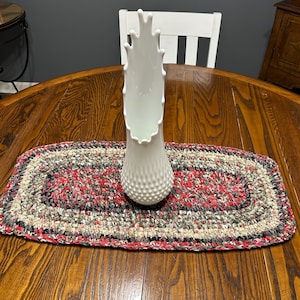 May include: A white, textured vase with a unique, organic shape stands on a handmade, oval-shaped table runner. The runner features a mix of red, gray, and beige yarns. The vase is approximately 12 inches tall.