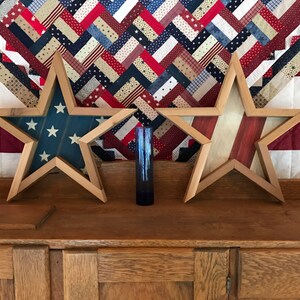 May include: Two wooden star-shaped frames with an American flag design. The frames are displayed on a wooden cabinet with a red, white and blue quilt in the background.