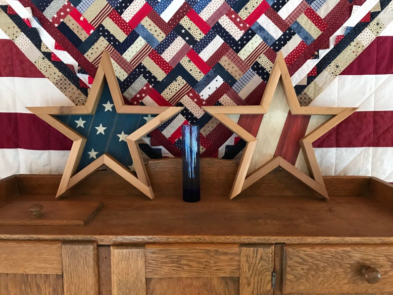 May include: Two wooden star-shaped frames with an American flag design. The frames are displayed on a wooden cabinet with a red, white and blue quilt in the background.