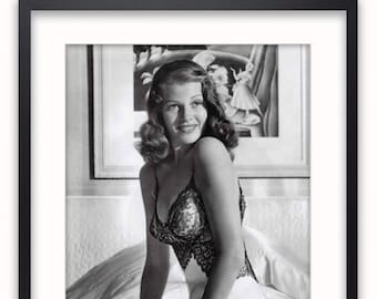 May include: A black and white photo of a woman in a satin slip dress and lace lingerie. She is sitting on a bed with a painting in the background.