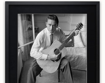 May include: Black and white photo of Johnny Cash sitting on a chair, holding an acoustic guitar. The photo is framed in a black frame with a white mat. The text "JOHNNY CASH" is printed below the photo.