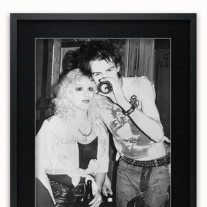 May include: Black and white photograph of a couple, a woman with blonde hair and a man with dark hair, standing close together. The woman is wearing a white shirt and a black leather jacket. The man is wearing a white t-shirt with a graphic design and a black leather jacket. They are both holding bottles of beer.