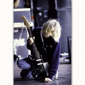 May include: A black electric guitar with a white pickguard and a sticker that says "Beauty is in a copycat" is held by a person with long blonde hair wearing a blue sweater and blue jeans. The person is kneeling on a gray floor.