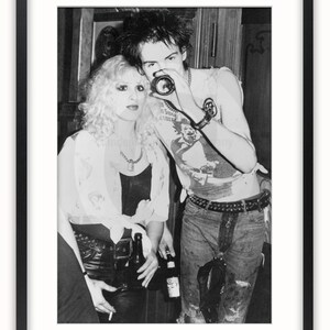 May include: A black and white photo of a man and a woman. The man is wearing a sleeveless shirt with a graphic on it and the woman is wearing a white shirt with a necklace. The man is holding a camera up to his eye and the woman is holding a cigarette in her hand.
