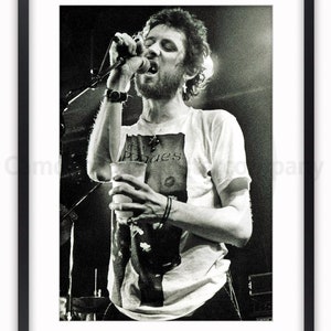 K&ouml;nnte beinhalten: Schwarz-wei&szlig; gerahmtes Poster einer Live-Musik-Performance. Eine Person singt in ein Mikrofon, h&auml;lt einen Becher und tr&auml;gt ein T-Shirt mit dem Aufdruck "Pogues". Das Bild ist eine Nahaufnahme des Darstellers auf der B&uuml;hne.