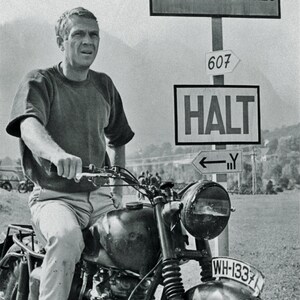 May include: Black and white image of Steve McQueen on a vintage motorcycle. The motorcycle has a headlight, a license plate, and a dark fuel tank. Signs in German read "Achtung" and "Schweizer Grenze". The film title "The Great Escape" is at the bottom.