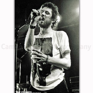 K&ouml;nnte beinhalten: Schwarz-wei&szlig; Konzertposter mit Shane MacGowan von The Pogues beim Singen in ein Mikrofon. Er tr&auml;gt ein wei&szlig;es T-Shirt mit Bandgrafik und h&auml;lt einen Becher. Der Bandname und Shanes Name sind unten aufgedruckt.