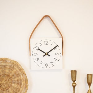 Puede incluir: Un reloj cuadrado blanco con números y manecillas negros que cuelga de una correa de cuero marrón. El reloj está montado en una pared blanca.