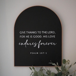 May include: Black arched wooden sign with white text that reads "GIVE THANKS TO THE LORD, FOR HE IS GOOD; HIS LOVE endures forever PSALM 107:1".
