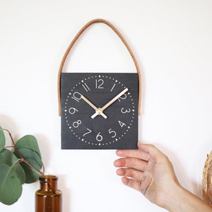 May include: A black square clock with white numbers and a gold-colored hour and minute hand. The clock has a brown leather strap for hanging.