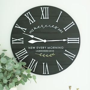 May include: Black wooden clock with white Roman numeral markings and white hands. The clock face has the text "His mercies are new every morning" and "Lamentations 3:22-23" in white lettering.