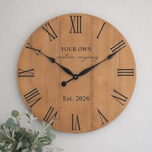 May include: A round wooden wall clock with a light brown finish and vertical plank detailing. It features black Roman numerals, black metal hands, and the printed text "YOUR OWN custom saying" and "Est. 2026" in the center of the clock face.