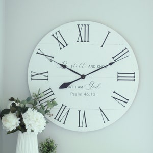 May include: White farmhouse style clock with black Roman numeral hour markers and black hands. The clock face has the text "Be still and know that I am God, Psalm 46:10".