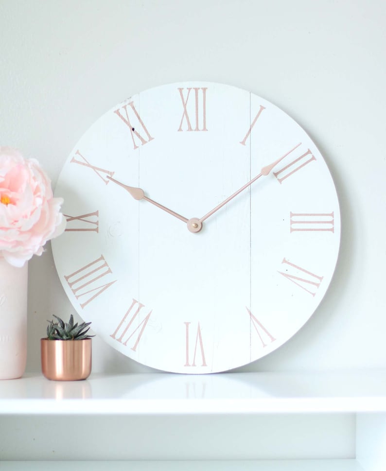May include: White farmhouse style clock with roman numerals in a rose gold color. The clock hands are also rose gold.