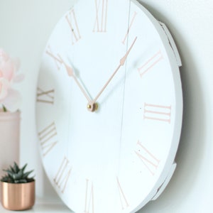 May include: White round clock with roman numerals in rose gold. The clock hands are also rose gold.