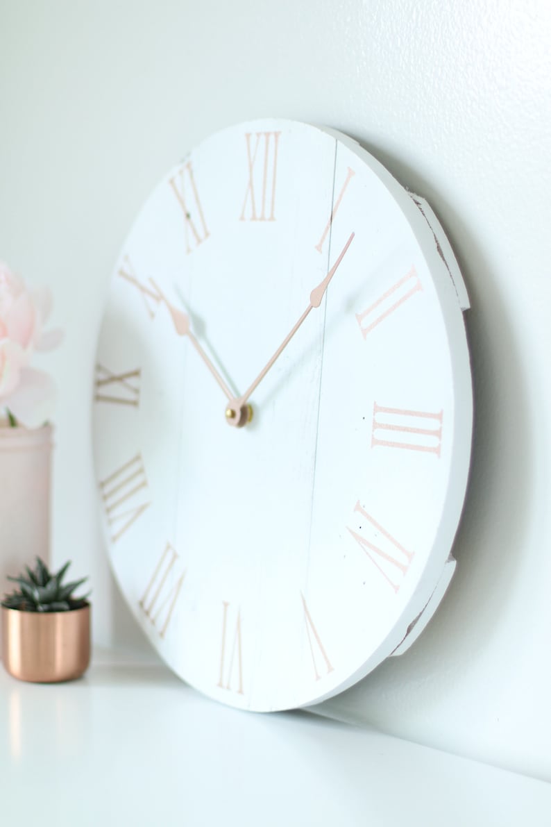 May include: White round clock with roman numerals in rose gold. The clock hands are also rose gold.